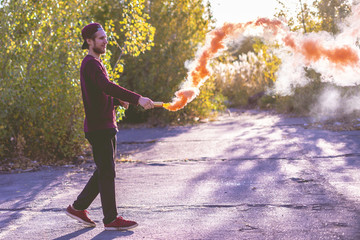 young happy male hipster run jump and explode the orange smoke bomb on a summer day in the green...