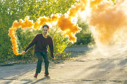 young happy male hipster run jump and explode the orange smoke bomb on a summer day in the green park f