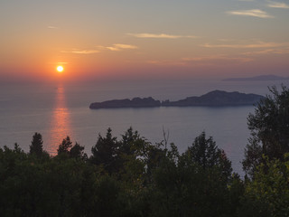 Beautiful colorful orange sunset sun falling to the ocean with rocks in mediterranean sea on Porto Timony, green bush in foreground Corfu,island, Greece