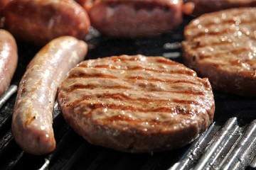 Shallow focus close up of tasty assorted delicious sausages and burgers sizzling and cooking on a barbecue griddle plate, outside in bright summer sunshine