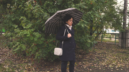 A young woman stands with a dark umbrella. Against a green tree. Autumn weather. Leaf litter. Brown. Dark coat and leather boots. The wind blows.