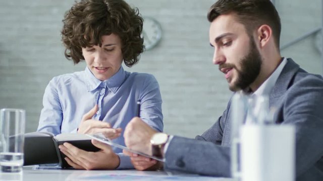Medium shot of two colleagues, man and woman, discussing project at office desk, woman using notebook for planning and man adding notes to document attached to clipboard