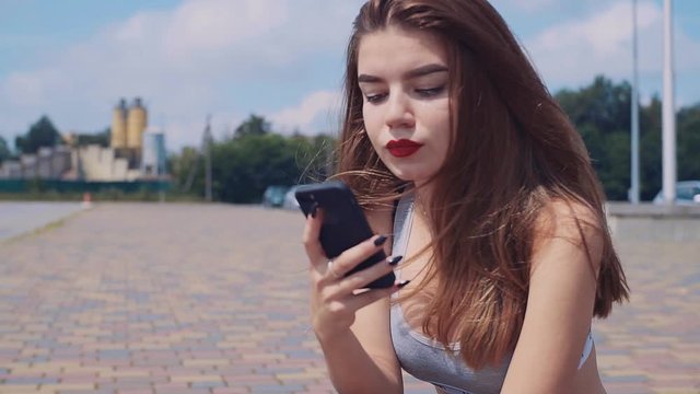 Brown-haired young Caucasian woman holding smartphone in hand, looking attentively at screen. Portrait of stunning Slavic student. Outdoors. Good weather.