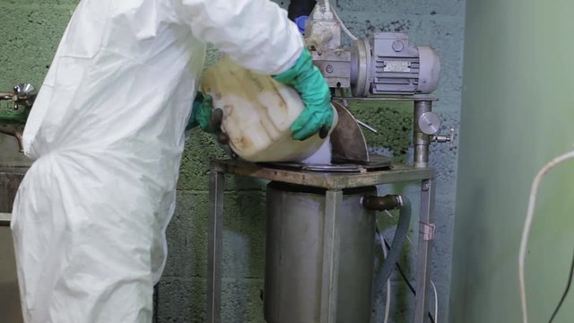 Lab worker wearing dress-uniform and green gloves is pouring white powder from large tray into big steel tank placed in corner of factory basement with dimmed light.