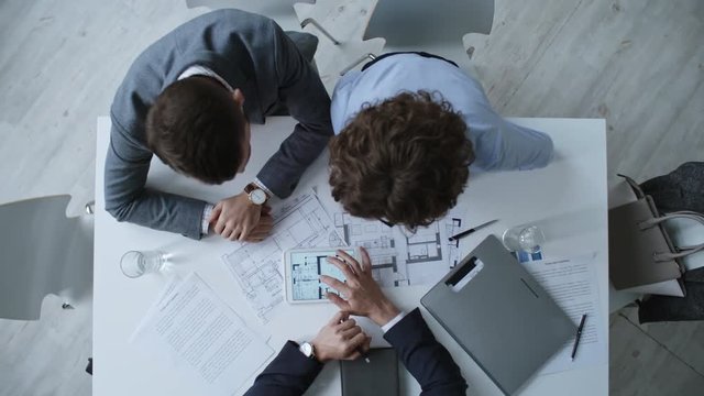 Zoom out shot of man and woman looking at apartment blueprints when sitting at desk in real estate agency, top view
