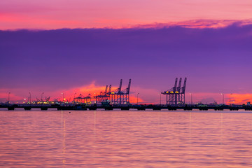 Fototapeta premium Logistics and transportation of Container Cargo with working crane bridge in shipyard. Cargo shipping and commercial terminal in seaport at sunset. Industrial landscape with gantry cranes in sea port.
