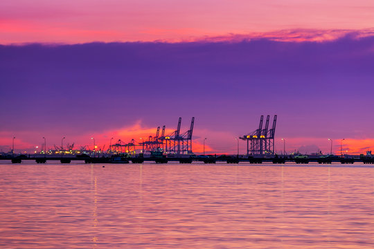 Logistics and transportation of Container Cargo with working crane bridge in shipyard. Cargo shipping and commercial terminal in seaport at sunset. Industrial landscape with gantry cranes in sea port.