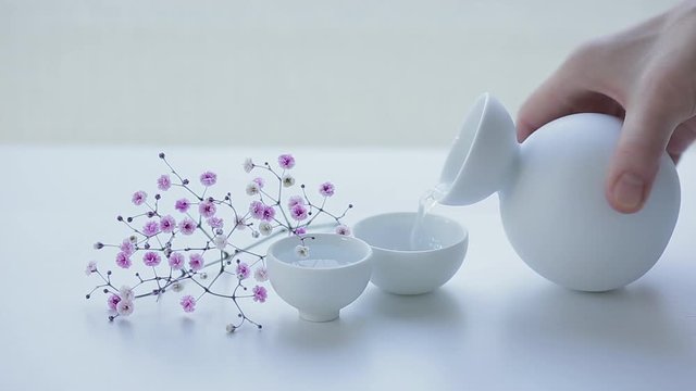 CINEMAGRAPH - seamless loop. Japanese sake is poured into ceramic glass from jug on white table, decorated with pink small flowers.