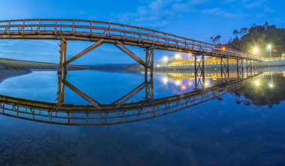 Obraz premium Amazing landscape at twilight in Buchupureo, a below view of the bridge that cross the river to access the beach and its reflection on the water illuminated by the blue hour and the first lamps, Chile