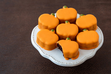 White platter with small orange frosted pumpkin cakes, one with bite taken out, on brown background

