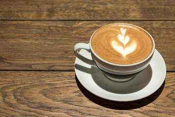 A white cup of cappuccino with a leaf shape on the top over a wooden background.