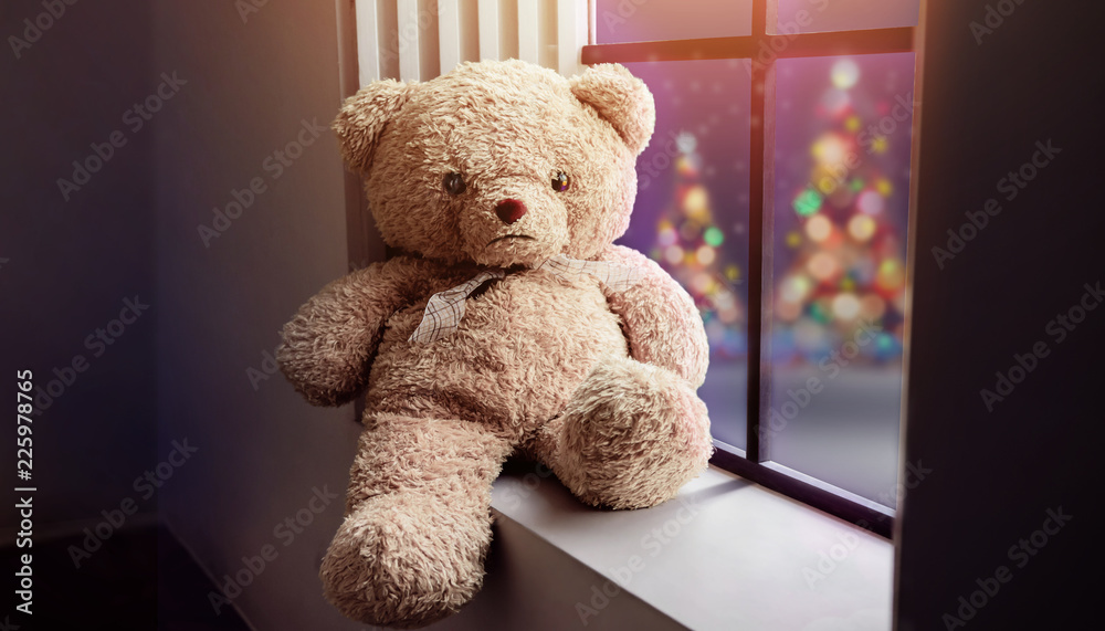 Wall mural Lonely Teddy Bear Sitting nearby Window in House on Christmas Night, Xmas Tree with Lights as outside view