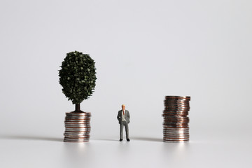 Miniature man standing between a pile of coins and a tree on a pile of coins.