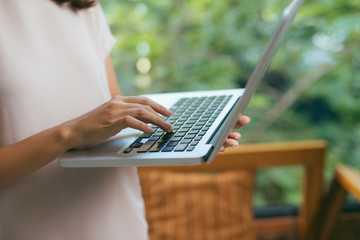 Portrait of young woman working holding laptop standing against panoramic window with garden view