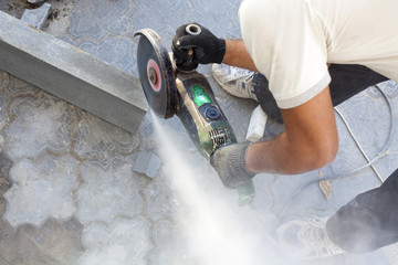 A worker at the construction site saws off a piece of concrete curb with angle grinder, circular electric saw, a tool in the hands of a worker, a sidewalk on a city street background close up - obrazy, fototapety, plakaty