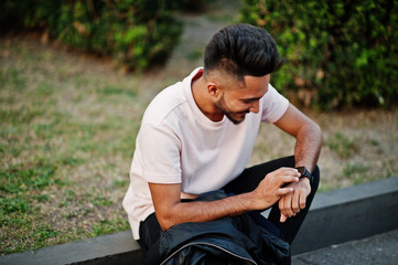 Stylish indian beard man at pink t-shirt. India model sitting at green garden of city and looking at his modern watches.