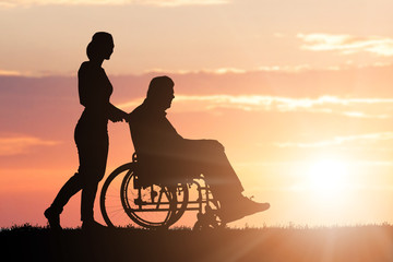 Fototapeta na wymiar Silhouette Of Woman Assisting Her Disabled Father On Wheelchair