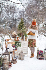Cozy mood of Christmas and New Year. A very beautiful girl in a white sweater and a red hat is surrounded by decor for the festive. Holds a basket with cones. Xmas wreath, forest with snow, lamp, fir.