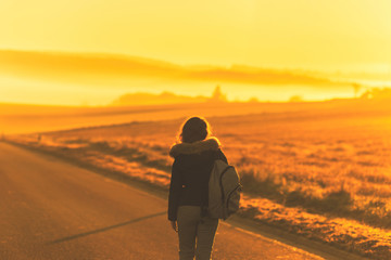 Fototapeta na wymiar Young woman in the winter jacket with a backpack on the road against the background of the sunrise autumn field. Travel and tourism concept