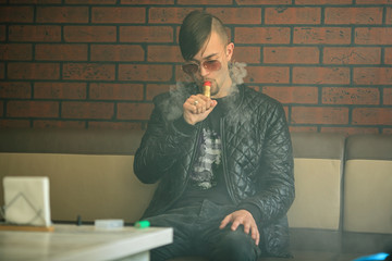 Vaping teenager. Young handsome bearded white man in a black jacket and sunglasses is smoking an...
