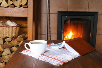 White mug of tea on the table in front of the fireplace with burning firewood.