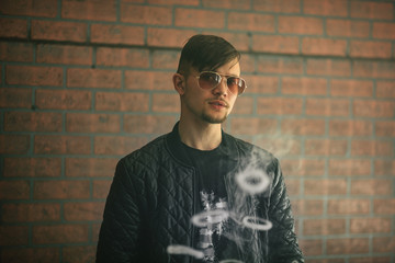 Vaping teenager. Young handsome bearded white man in a black jacket and sunglasses is smoking an...