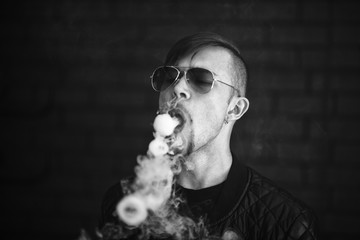 Vaping teenager. Young handsome bearded white man in a black jacket and sunglasses is smoking an...