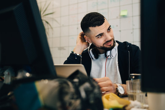 Confused young male computer programmer scratching head while sitting with laptop at creative office