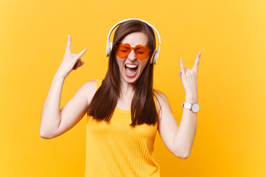 Portrait of excited fun girl in orange glasses listening music in headphones showing heavy metal rock sign, copy space isolated on yellow background. People sincere emotions concept. Advertising area.
