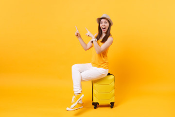 Traveler tourist woman in summer casual clothes, hat sit on suitcase isolated on yellow orange background. Female passenger traveling abroad to travel on weekends getaway. Air flight journey concept.