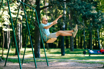 Odd adult bald man in sunglasses jumping from swing on ground in motion.  Childhood behavior....