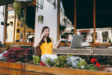 Happy girl in outdoors street cafe sitting at table with laptop pc computer, talking on mobile phone, conducting pleasant conversation, in restaurant during free time. Mobile office freelance concept