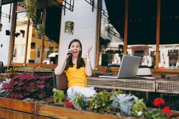 Happy girl in outdoors street cafe sitting at table with laptop pc computer, talking on mobile...