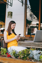 Happy smiling girl in outdoors street coffee shop cafe sitting at table with laptop pc computer, texting message on mobile phone friend, in restaurant during free time. Mobile office freelance concept