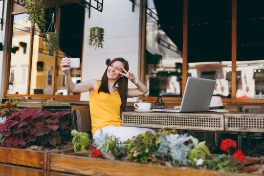 Happy smiling girl in outdoors street coffee shop cafe sitting at table with laptop pc computer doing taking selfie shot on mobile phone in restaurant during free time. Mobile office freelance concept