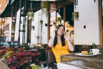 Fun woman in outdoors street coffee shop cafe sitting at table in yellow clothes listening music in headphones, relaxing in restaurant during free time. Mobile Office in summer. Lifestyle rest concept