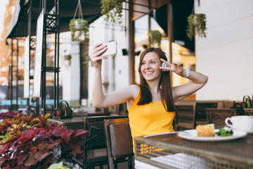 Attractive smiling woman in outdoors street coffee shop cafe sitting at table, listen music in headphones, doing selfie shot on mobile phone, relaxing in restaurant free time. Lifestyle rest concept.