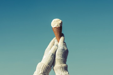 Female hand in knitted gloves with ice cream in a waffle horn on the blue sky background. Concept. 
