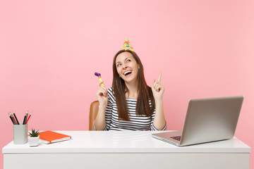 Laughing woman in birthday party hat with playing pipe celebrating while sit and work at white desk...