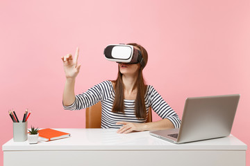 Girl in headset of virtual reality on head touch something like push on button or pointing at floating virtual screen work at desk with laptop isolated on pink background. Achievement business career.