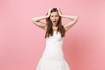 Portrait of irritated dissatisfied bride woman in white wedding dress standing screaming clinging to head isolated on pink pastel background. Wedding celebration concept. Copy space for advertisement.