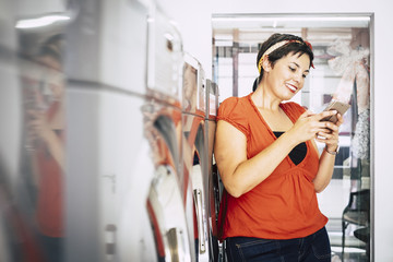 Happy beautiful caucasian middle age lady use smart mobile phone at the laundry mat automatic service shop while wait for her clothes washed and cleaned. Sharing on social media the activity