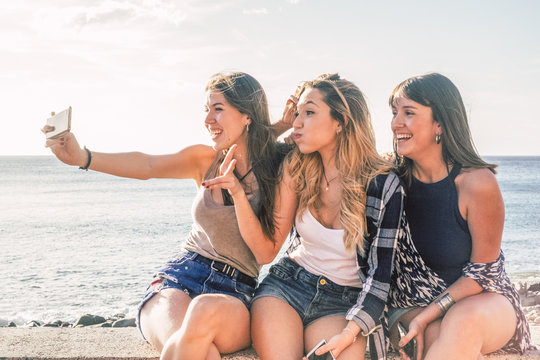 three young girls having fun together taking a picture selfie with a smart phone sit down with summer sea in background. friendship and funny day concept for beautiful women outdoor. cheerful females