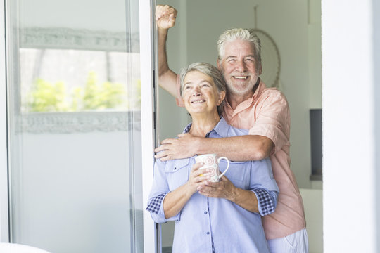 happy and beautiful retired couple of man and woman caucasian people enjoying the day together with love and joy at home loooking outside the door window. bright image with adult senior cheerful smile