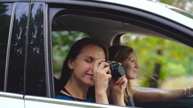 Two young women ride in a car and have fun. One of them takes a self photo on a film camera
