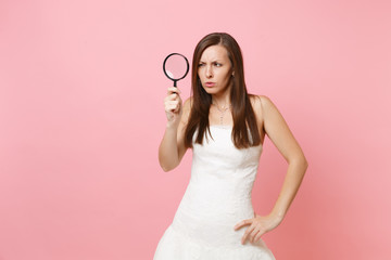 Portrait of suspicious bride woman in white wedding dress scrutinizing looking through magnifying glass isolated on pastel pink background. Wedding to do list. Organization of celebration. Copy space.