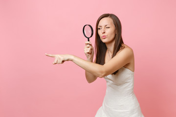 Concerned dissatisfied bride woman in wedding dress pointing index finger aside, looking through magnifying glass isolated on pastel pink background. Wedding to do list. Organization of celebration.