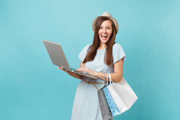 Portrait of young woman in summer dress, straw hat holding packages bags with purchases after online shopping, using laptop pc computer isolated on blue pastel background. Copy space for advertisement