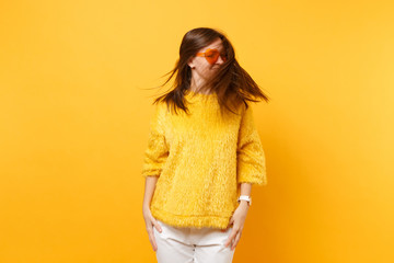 Funny young woman in fur sweater and heart orange glasses fooling around in studio with flowing hair isolated on bright yellow background. People sincere emotions, lifestyle concept. Advertising area.