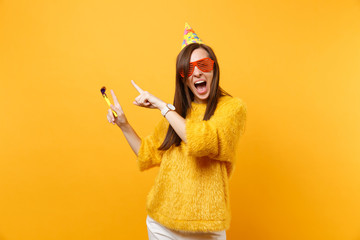 Excited happy woman in orange funny glasses, birthday hat with playing pipe pointing index fingers...